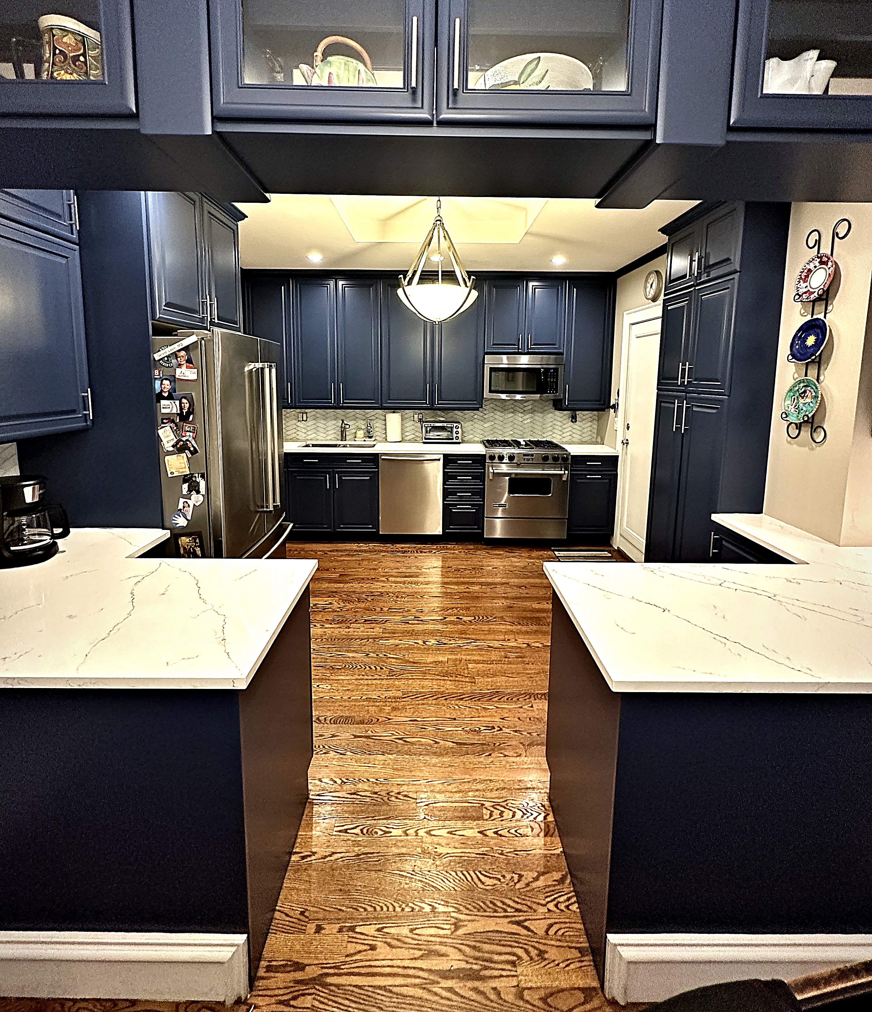 A kitchen with deep navy cabinets, glass upper doors, white stone countertops, and warm oak floors, refinished by GK Painting.